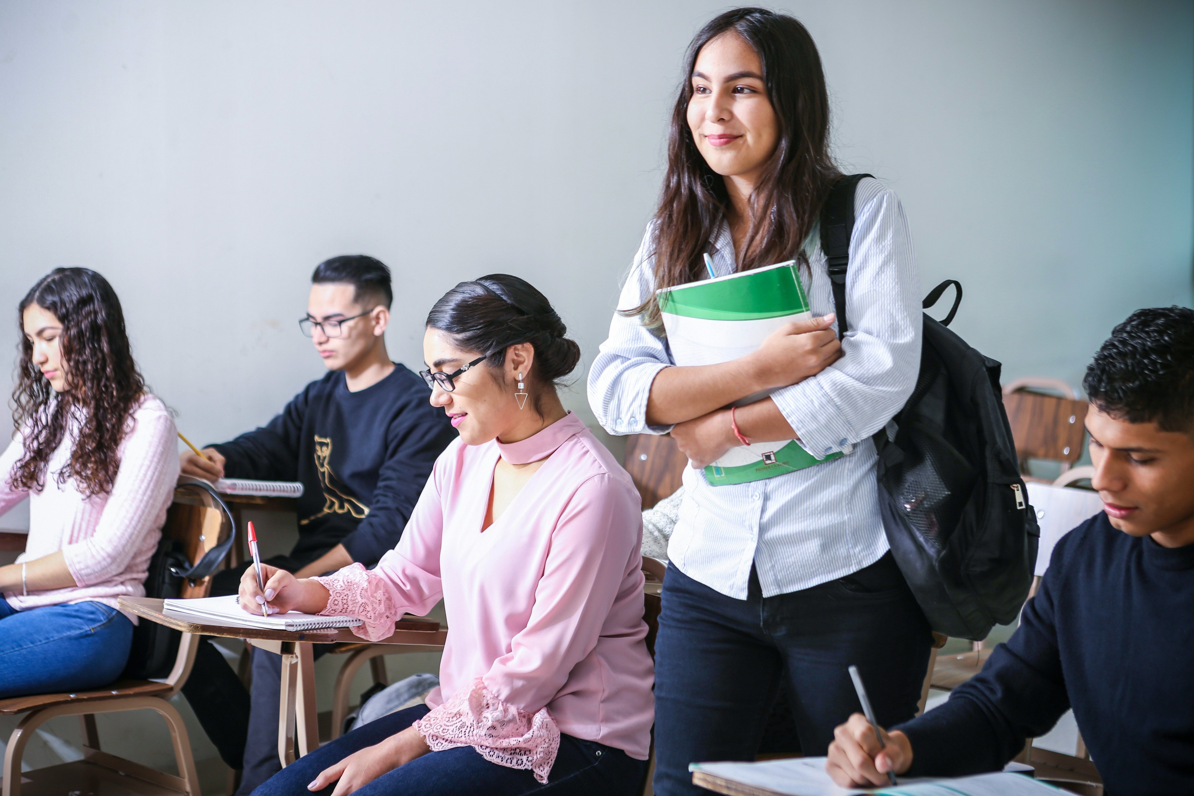 Students in a classroom, including a woman standing with a green and white textbook.