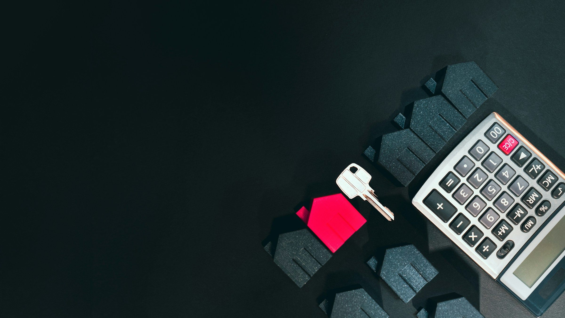 A silver house key, a small red house model and a calculator on a black surface
