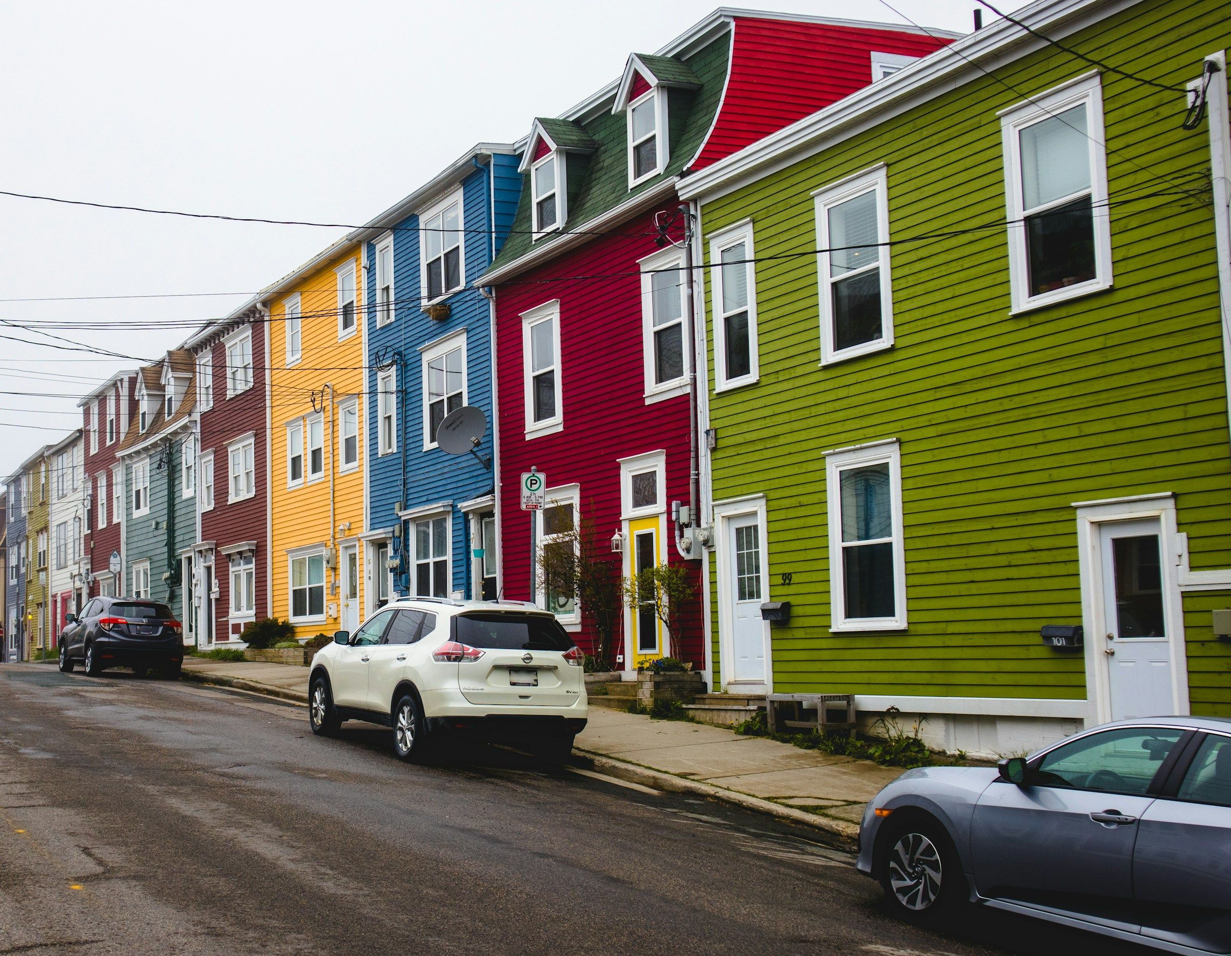 A row of brightly coloured attached houses on a steep street with cars parked along it