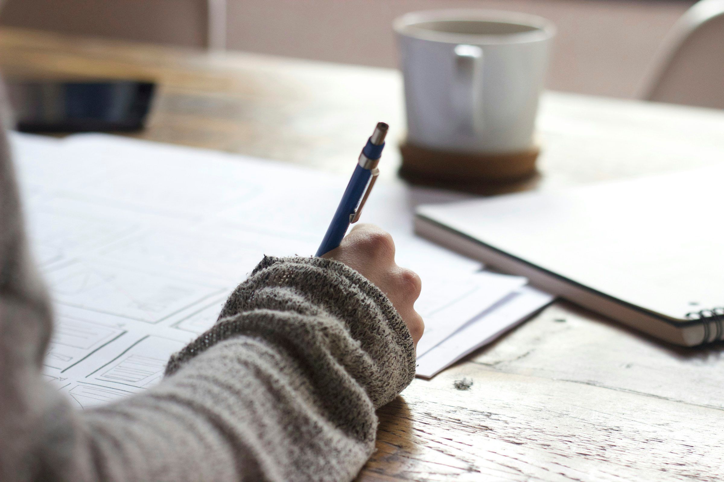 Person writing on a wooden table beside a white mug