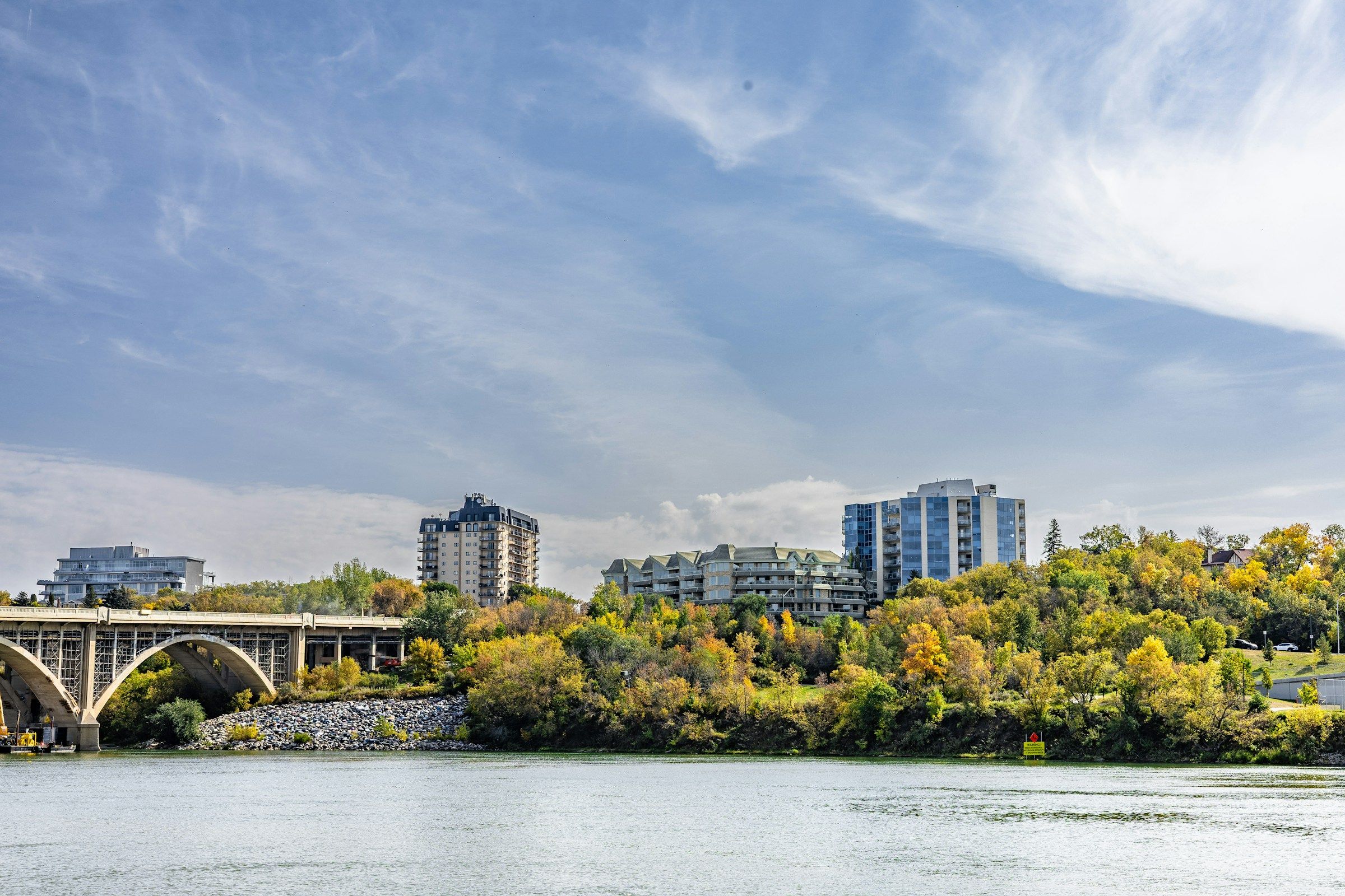 A city skyline behind a bridge over a river lined with autumn trees